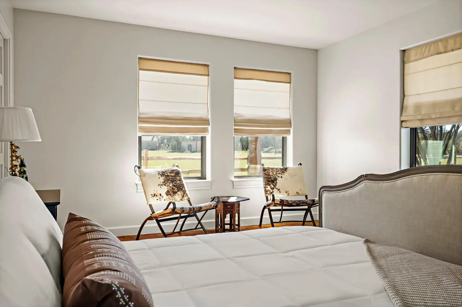 Dormitorio minimalista con una cama bien hecha, dos sillas, una mesa pequeña junto a la ventana y estores romanos de un suave tono beige que filtran la luz del sol.