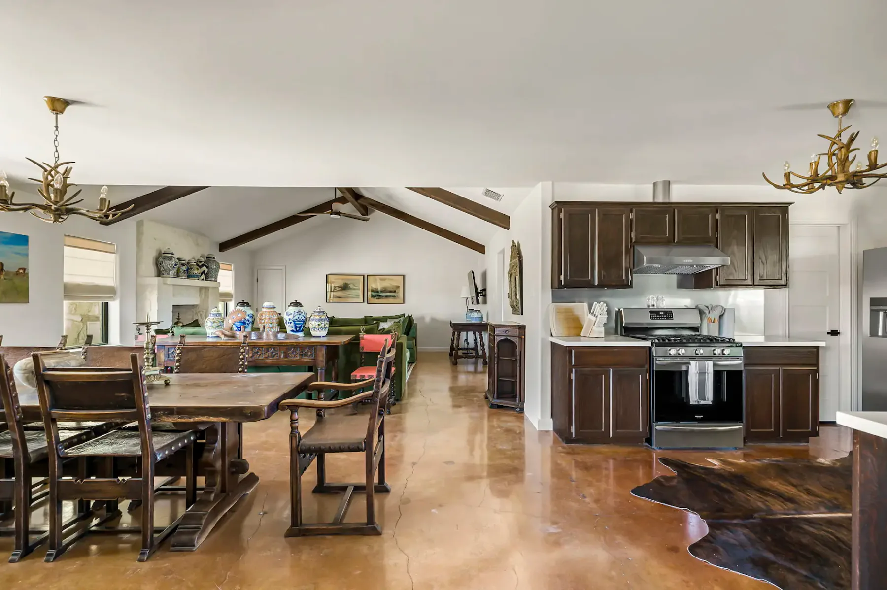Zona de estar espaciosa de concepto abierto con decoración rústica, una mesa de comedor de madera, armarios de cocina de madera oscura y lámparas de araña de astas.