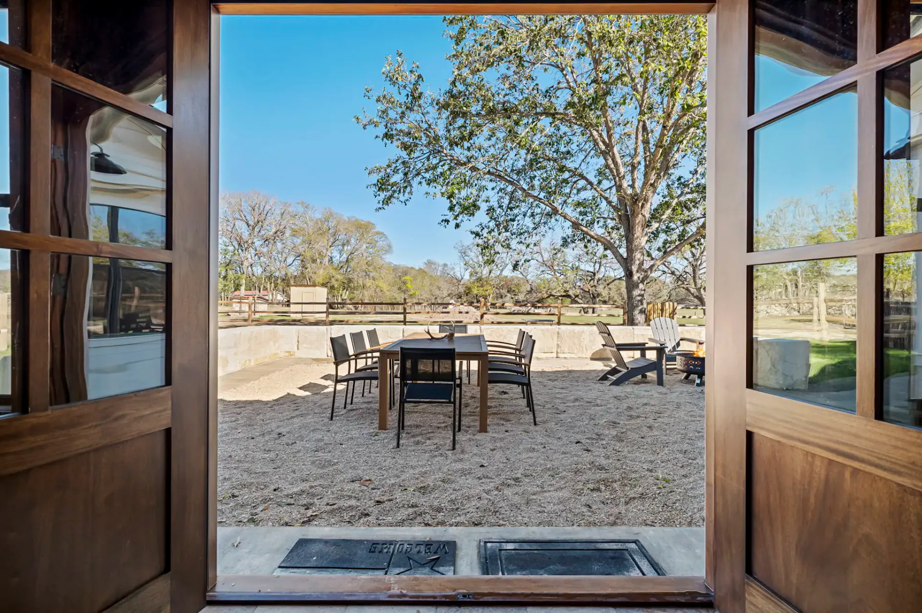 Vista a través de unas puertas de madera abiertas hacia un patio con mesa de comedor, sillas y un brasero, situado en un terreno arenoso con árboles grandes y un cielo azul despejado.