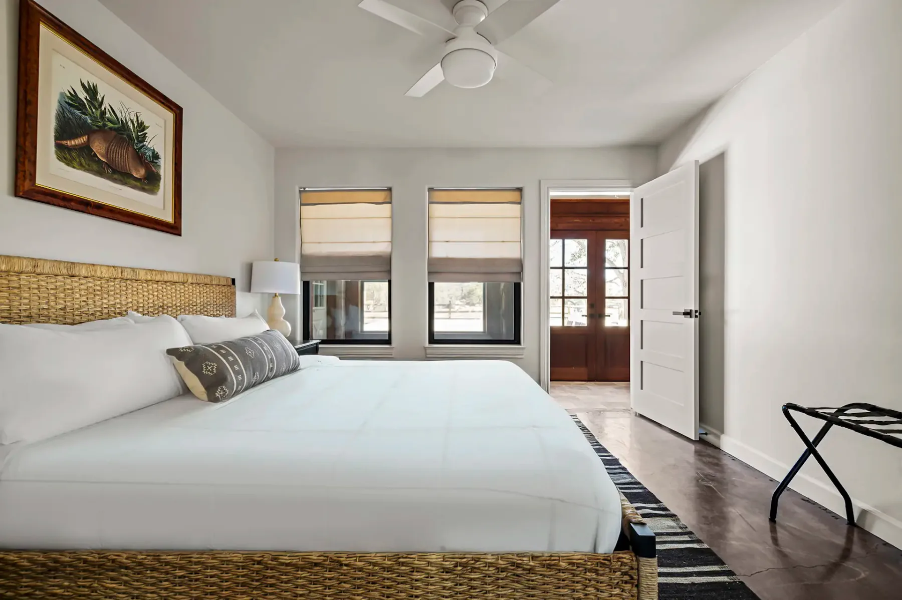 Dormitorio luminoso con una cama grande, cabecero de mimbre, dos ventanas, un ventilador de techo, una puerta abierta y obras de arte en la pared. Decoración minimalista y luz natural.