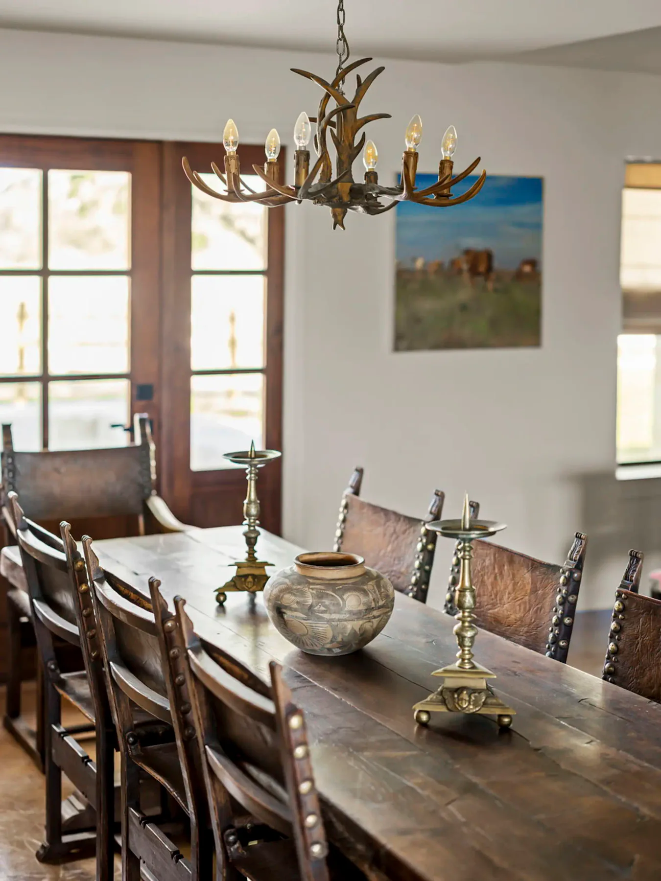 Comedor rústico con una larga mesa de madera rodeada de sillas de cuero, un jarrón decorativo y candelabros bajo una lámpara de araña de astas.