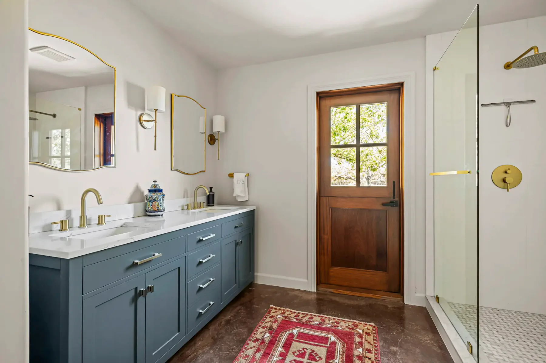 Baño moderno con mueble de lavabo azul, dos lavabos, grifería dorada, puerta de madera, ducha con cerramiento de vidrio y alfombra con estampado rojo sobre un suelo oscuro.