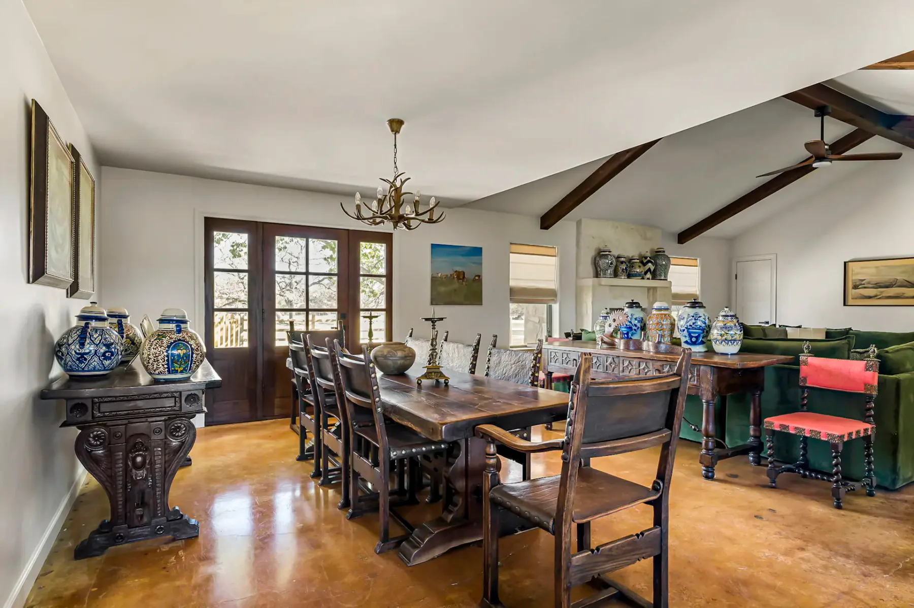 Comedor rústico con mesa y sillas de madera, lámpara de araña de astas, jarrones decorativos y grandes ventanas que permiten la entrada de luz natural.