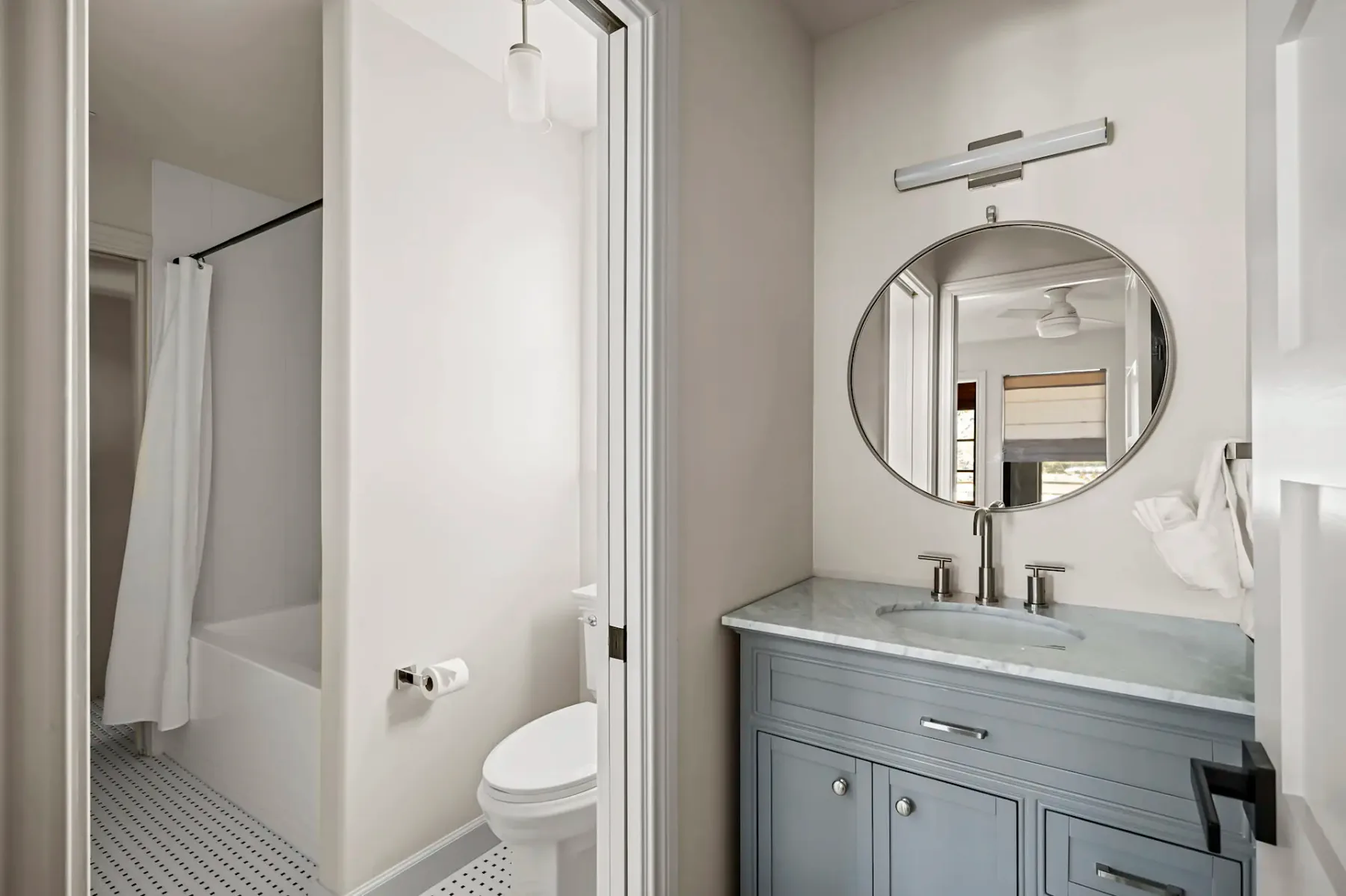 Baño moderno con un espejo redondo sobre un mueble de lavabo gris, encimera de mármol y elementos blancos. Al fondo se ve una bañera con cortina.