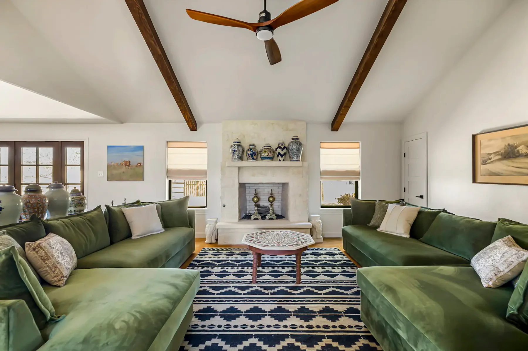 Sala de estar espaciosa con sofás verdes, una alfombra estampada, una mesa central de madera, techo con vigas y una repisa de chimenea revestida de azulejos decorativos.