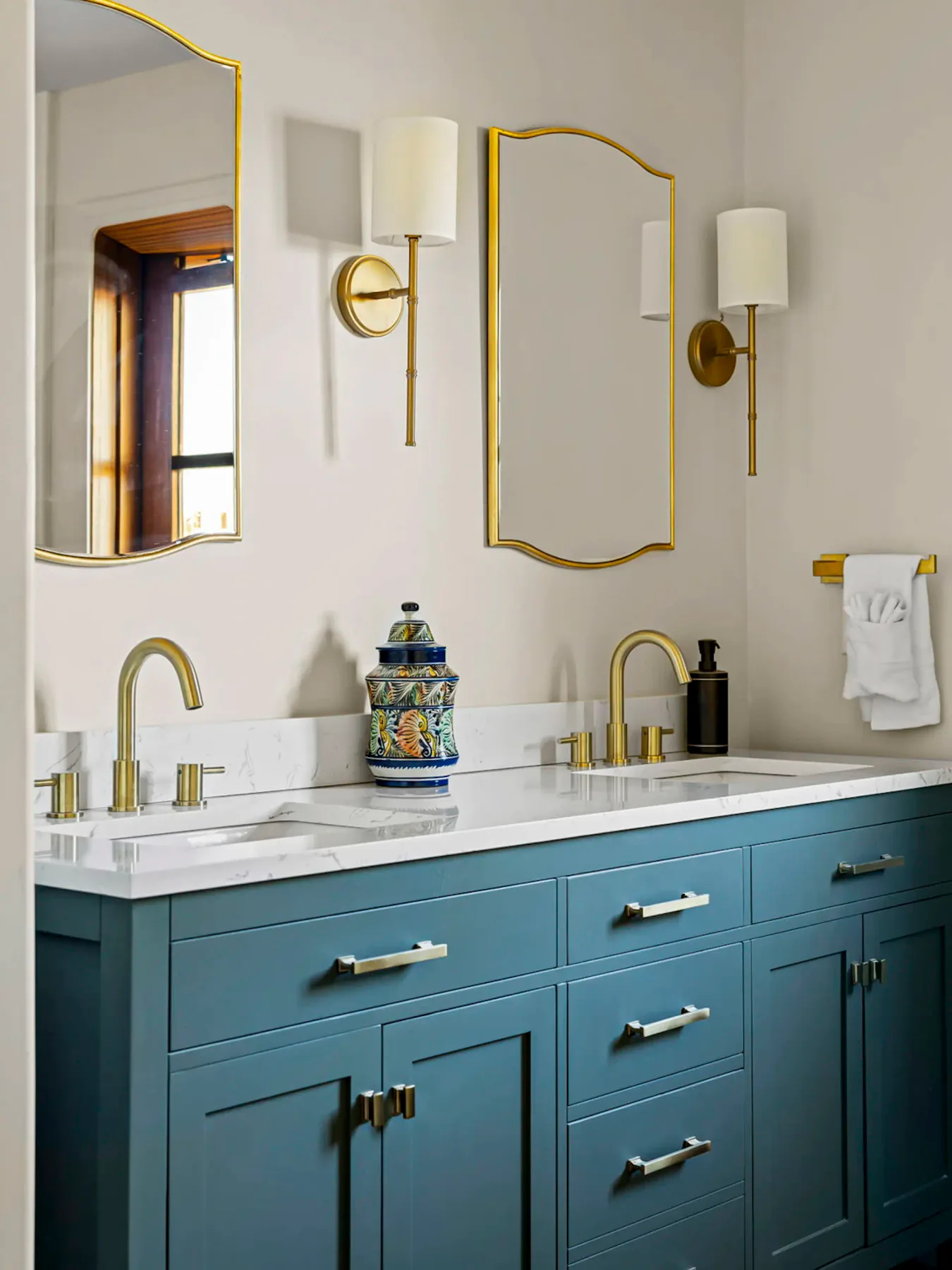 Baño con mueble de lavabo doble de color verde azulado, grifería dorada, dos espejos y apliques de pared. Hay un recipiente decorativo y una toalla sobre la encimera blanca.