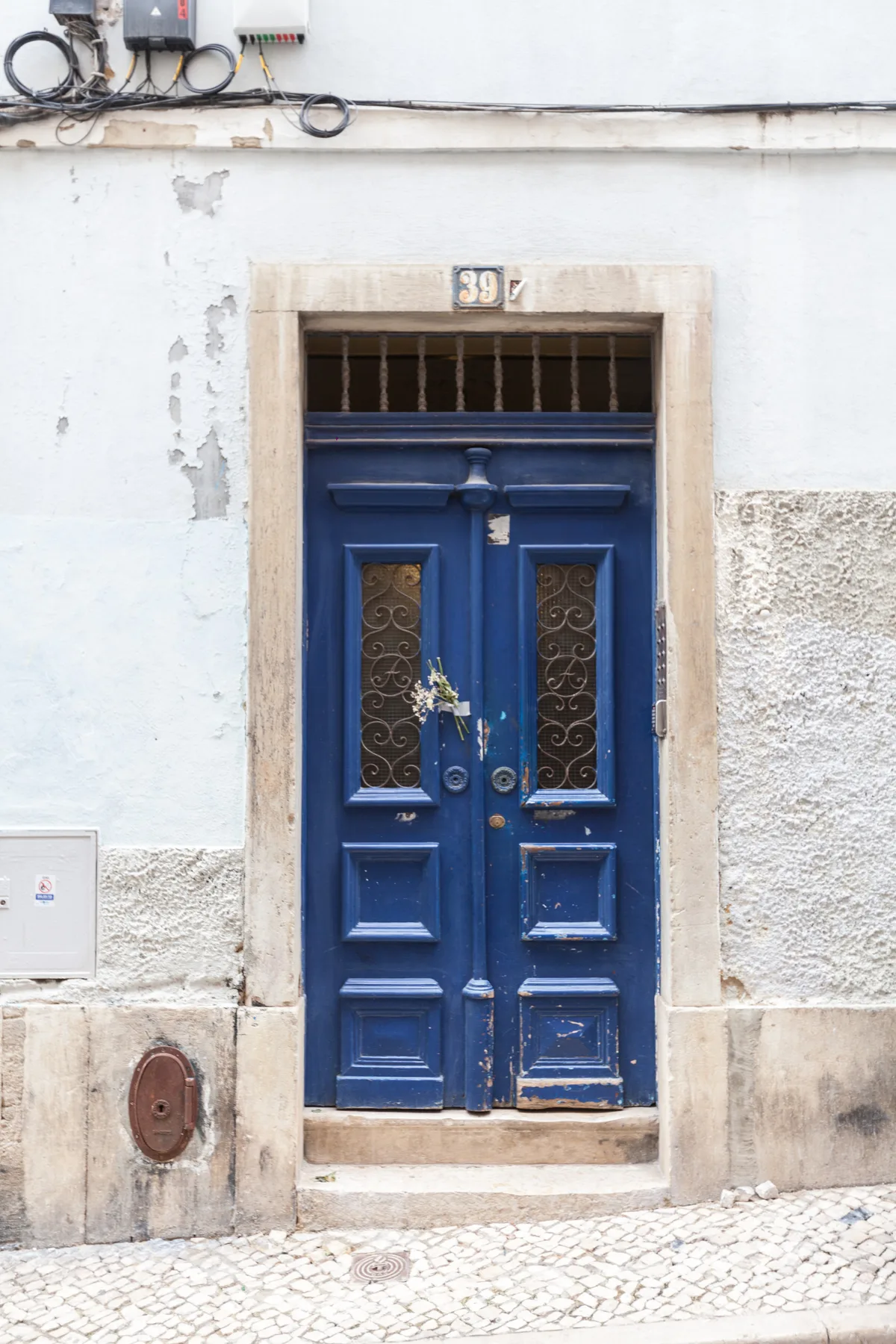 Una puerta doble azul en una pared exterior clara, sobre una acera empedrada.