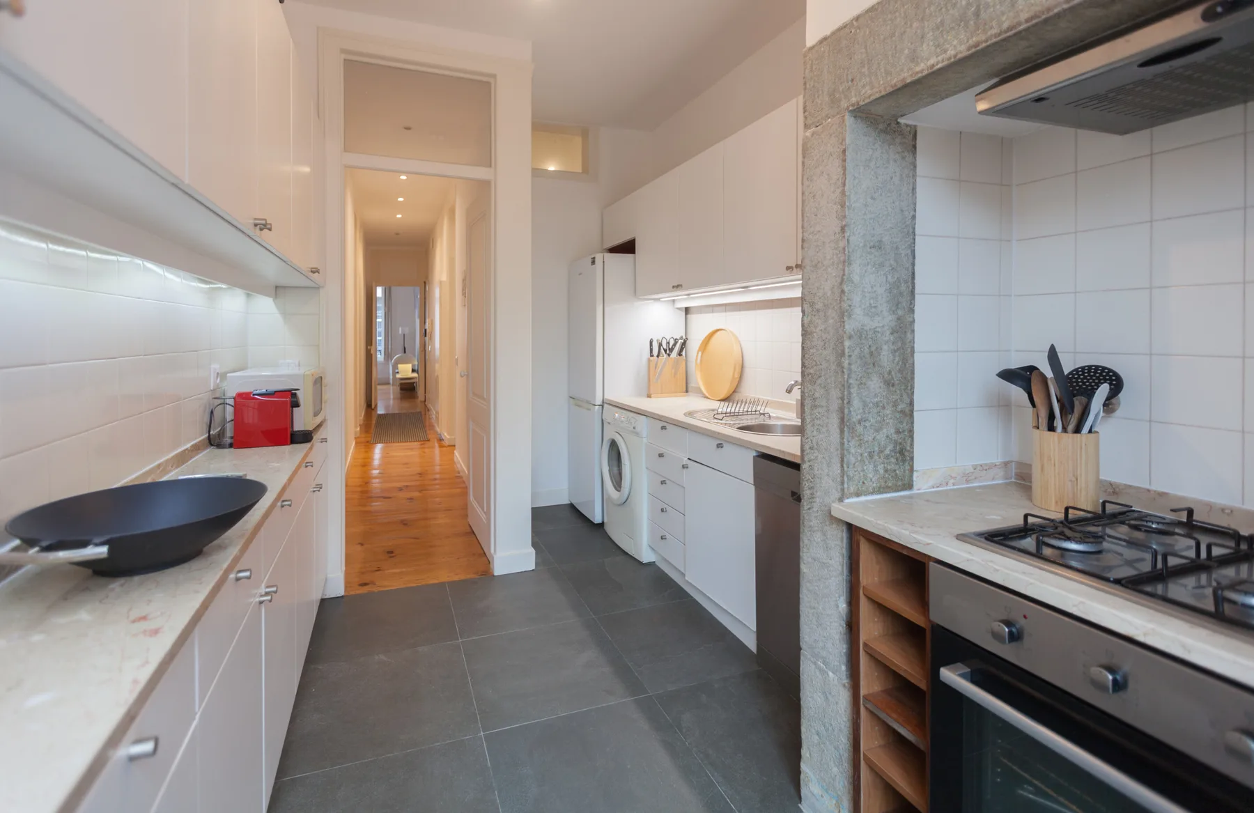 Una cocina alargada con armarios blancos, una placa de cocina y un pasillo al fondo.