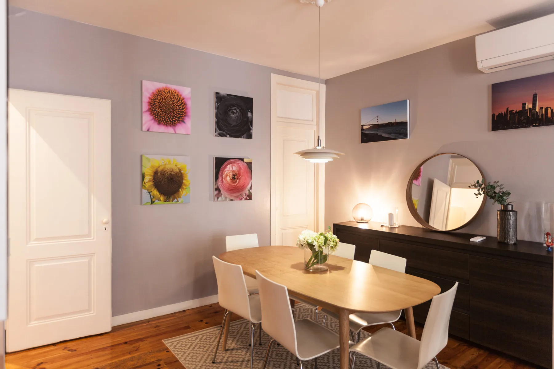 Comedor con mesa de madera, asientos blancos y cuadros coloridos en la pared.