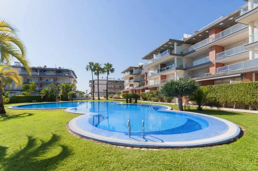 A curved swimming pool sits on a lawn beside apartment buildings.