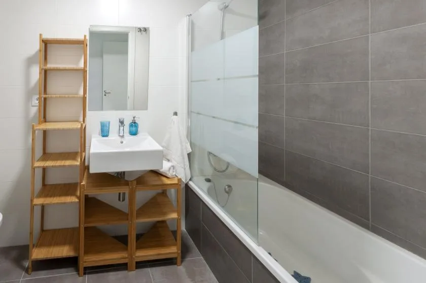A bathroom with a sink, wooden shelving, and a glass-screened bathtub.