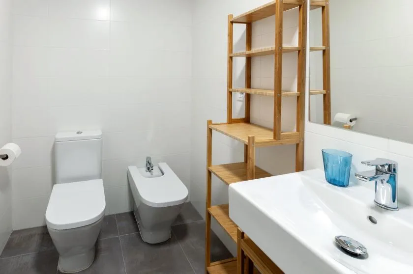 A bathroom with a toilet, bidet, sink, and wooden shelving.