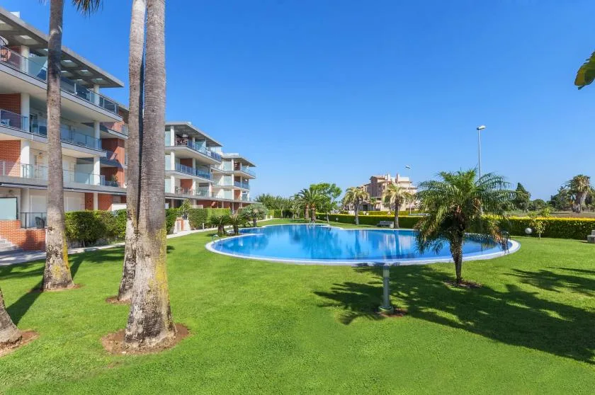 An outdoor swimming pool sits among green lawns and apartment buildings under a clear sky.