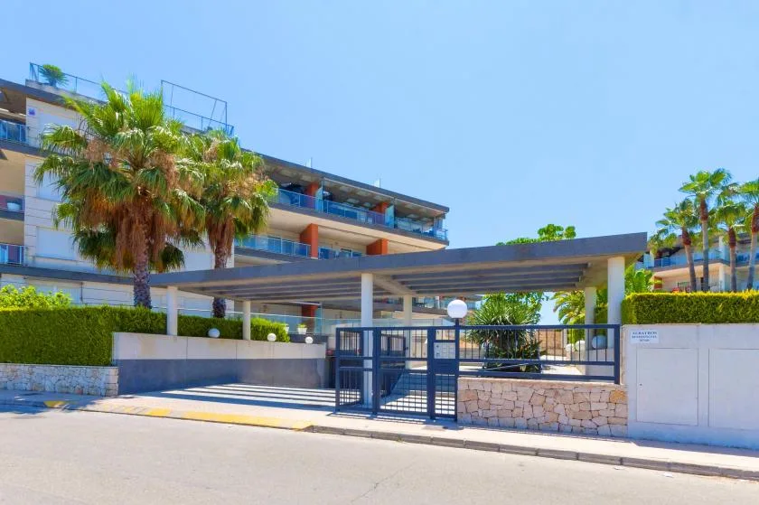 A modern apartment building stands behind a gated entrance and palm trees.