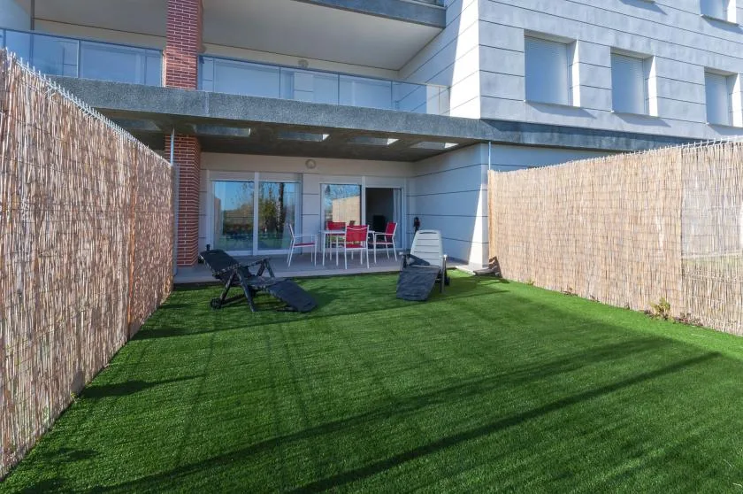 A fenced outdoor patio with green grass and seating beside a modern building.