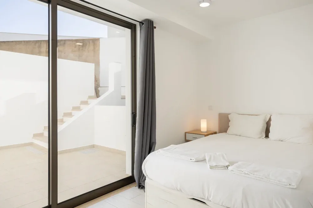 Un dormitorio luminoso con ropa de cama blanca junto a una gran puerta corrediza de vidrio.