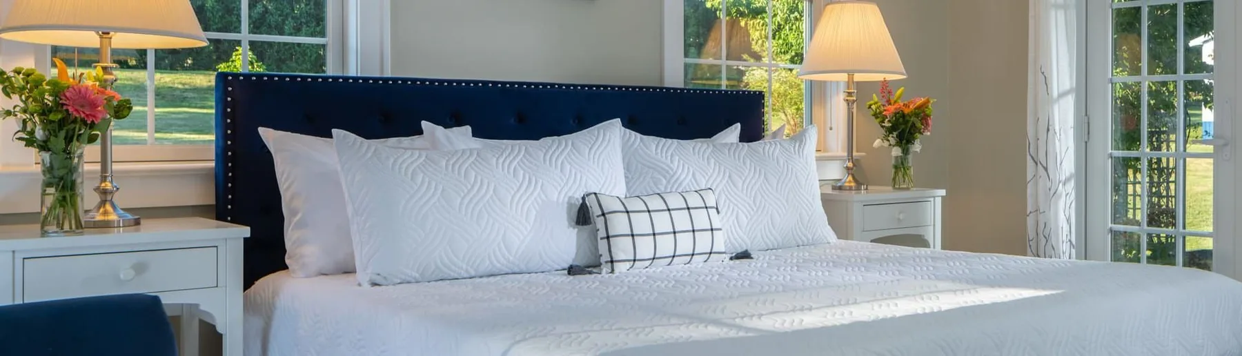 Cama con tapizado azul marino, ropa de cama blanca, mesitas de noche de madera, lámparas y ventanas