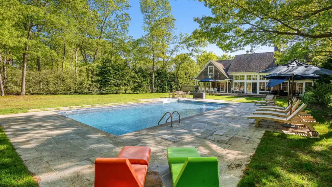 Una piscina al aire libre junto a una casa, con asientos de colores en el patio de piedra.