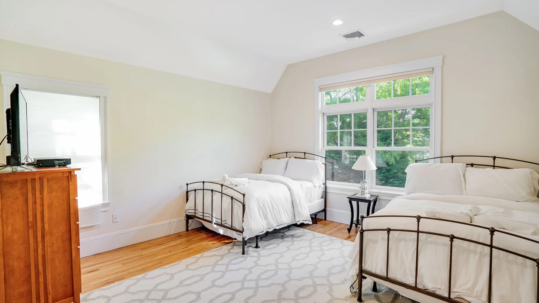 Un dormitorio luminoso con dos camas, ventanas grandes y una cómoda de madera.