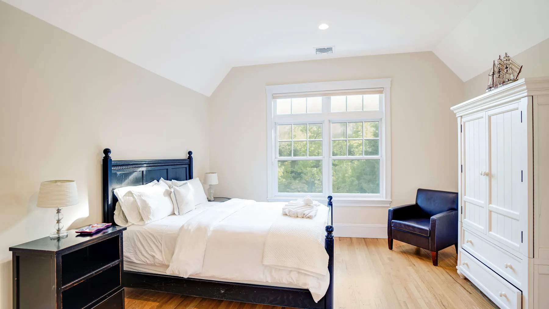 Un dormitorio luminoso con una cama, una ventana amplia y un sillón oscuro.