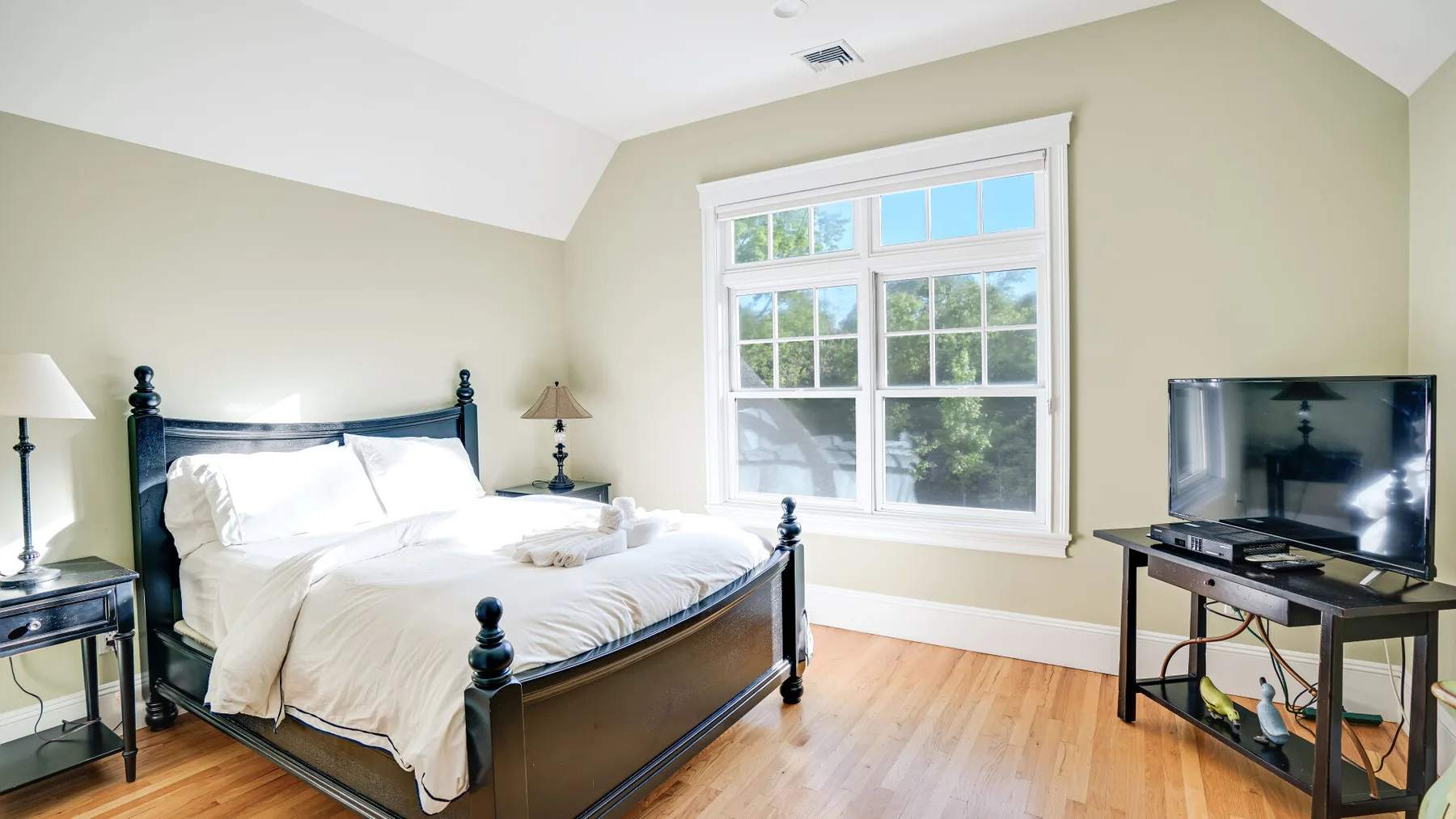 Un dormitorio luminoso con una cama oscura, ropa de cama blanca y un televisor junto a la ventana.