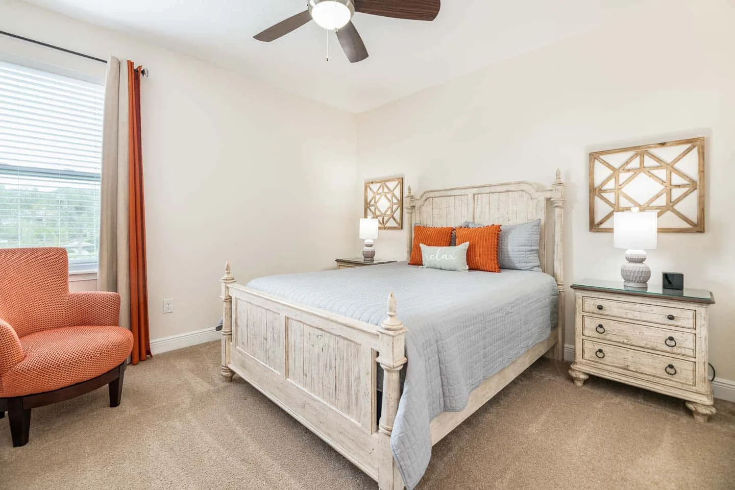 Un dormitorio con una cama, mesitas de noche y un sillón naranja junto a la ventana.