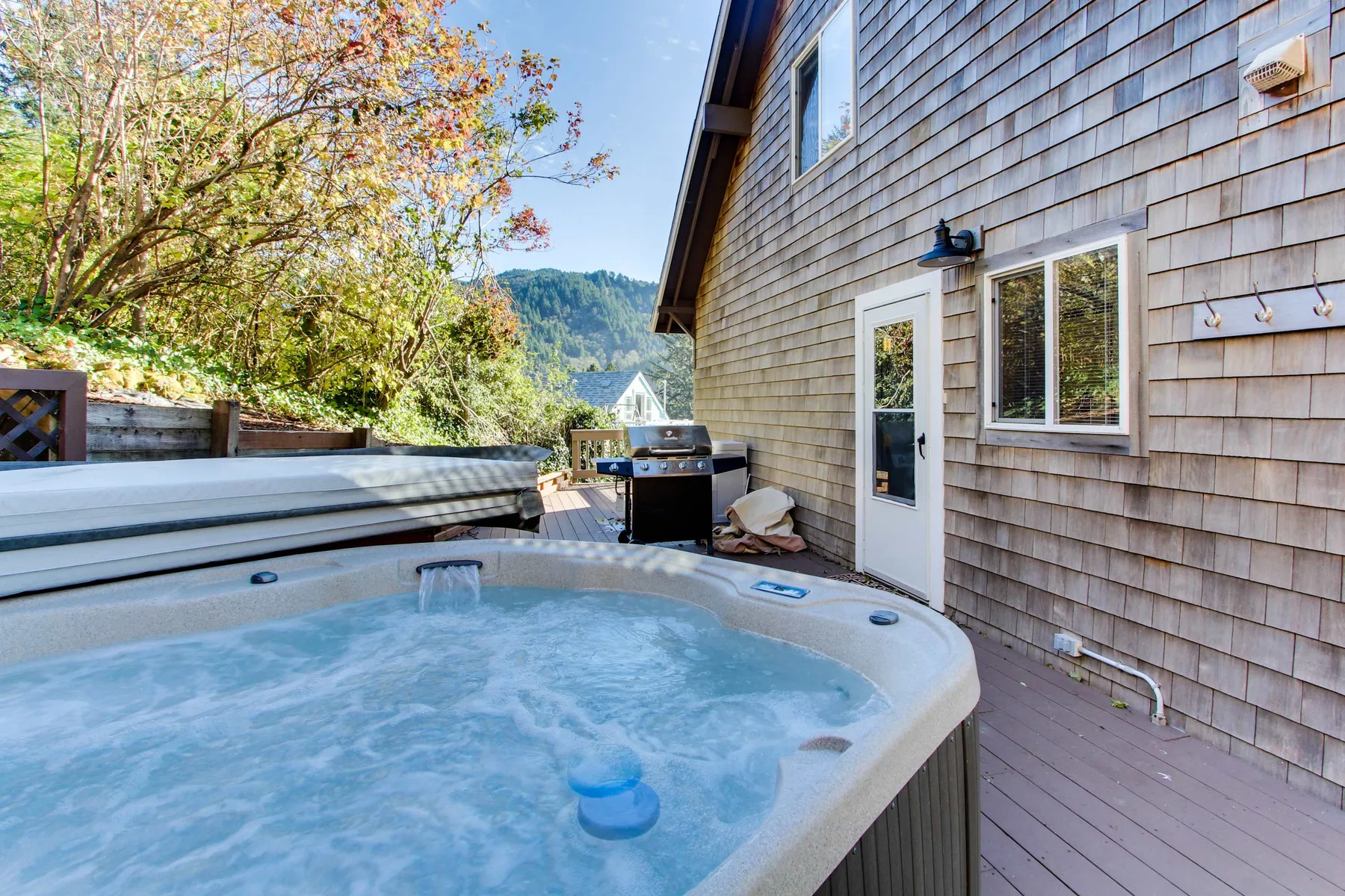 An outdoor hot tub sits beside a house deck, with trees and hills beyond.