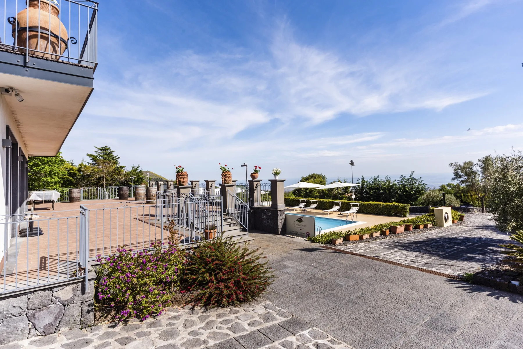 Una terraza exterior y una piscina dan a un jardín ajardinado bajo un cielo despejado.