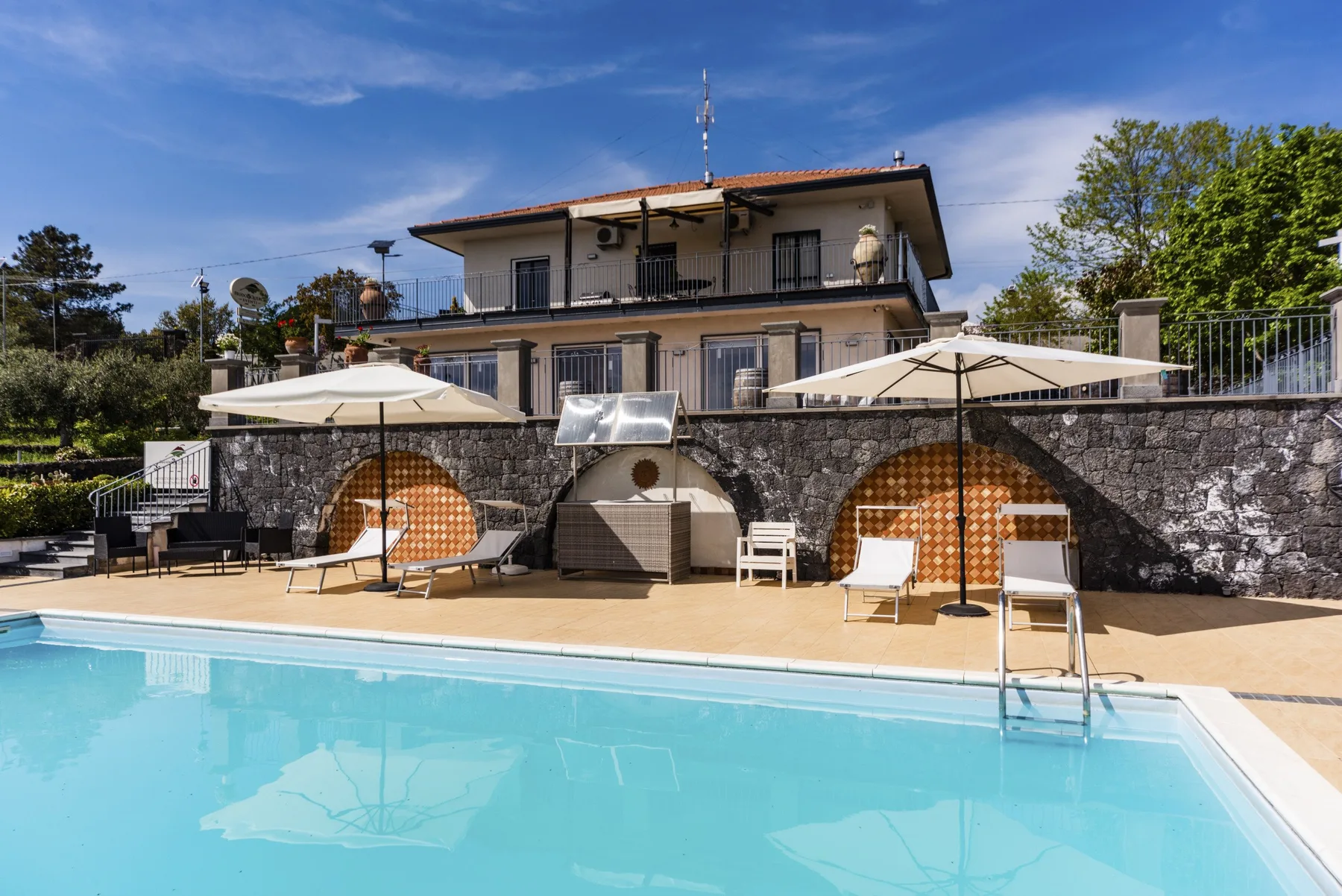 Una piscina junto a una terraza de piedra con sombrillas y una casa.