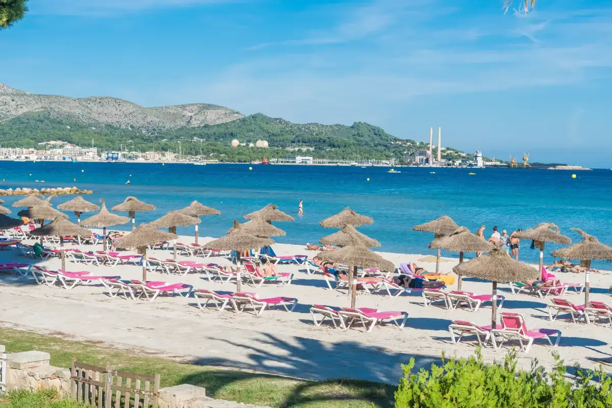 A sandy beach lined with straw parasols and loungers overlooks a blue bay.