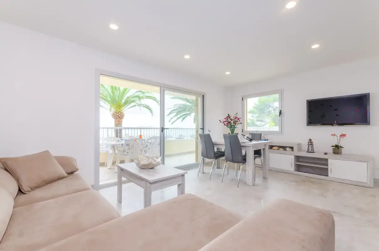 A bright living and dining area opens onto a balcony with palm trees outside.