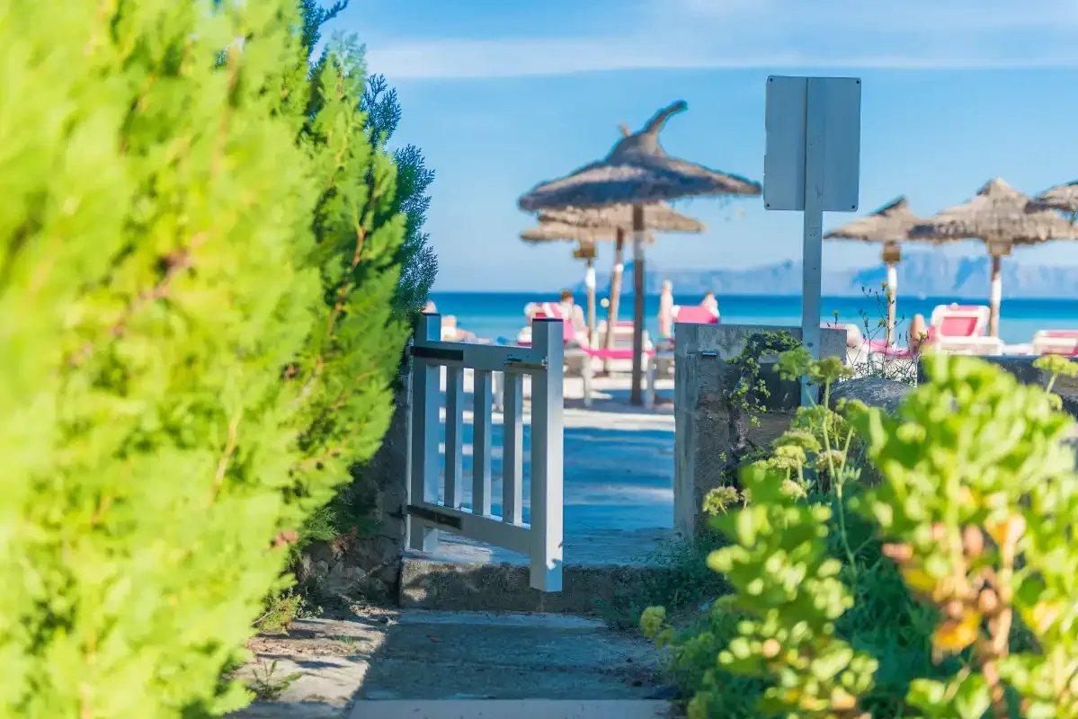 A seaside path leads past a small gate to a beach with umbrellas and loungers.