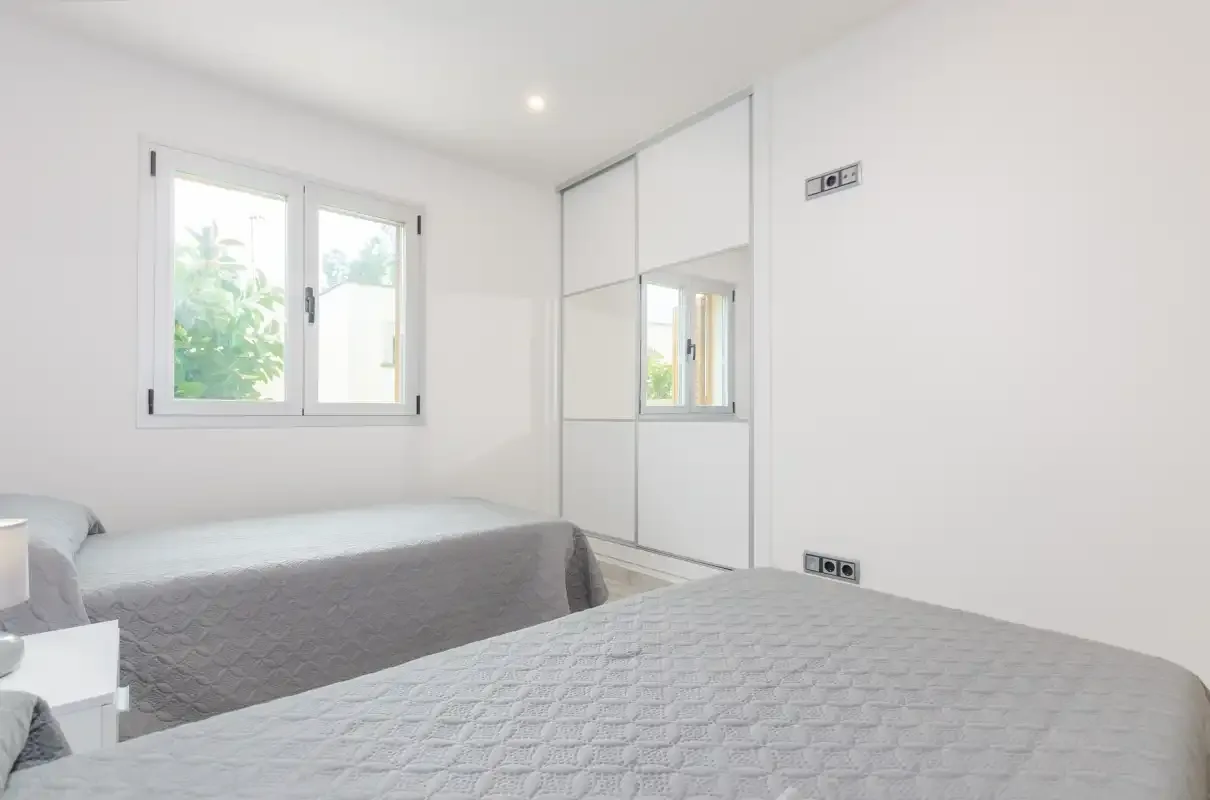 A bright bedroom with two beds and a window overlooking greenery.