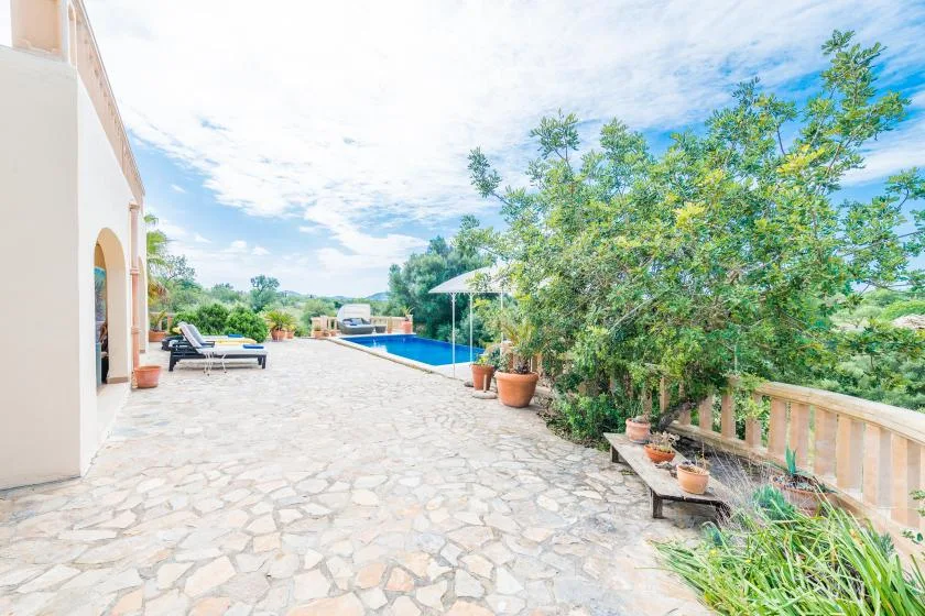 A sunlit stone patio beside a swimming pool, with outdoor seating and leafy plants.