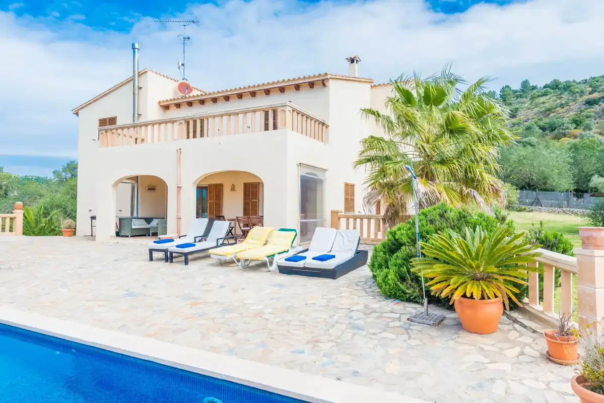 A sunlit villa patio with poolside loungers and palm trees.