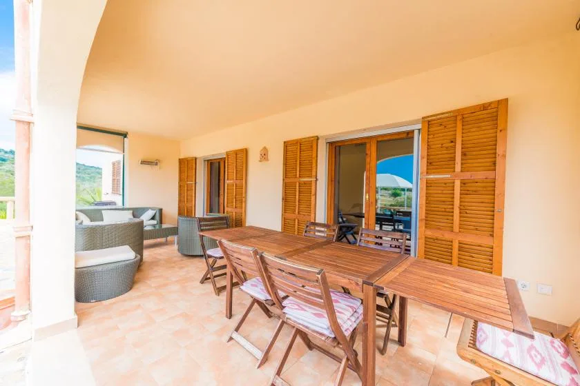 A covered patio with a wooden dining table and seating beside shuttered doors.
