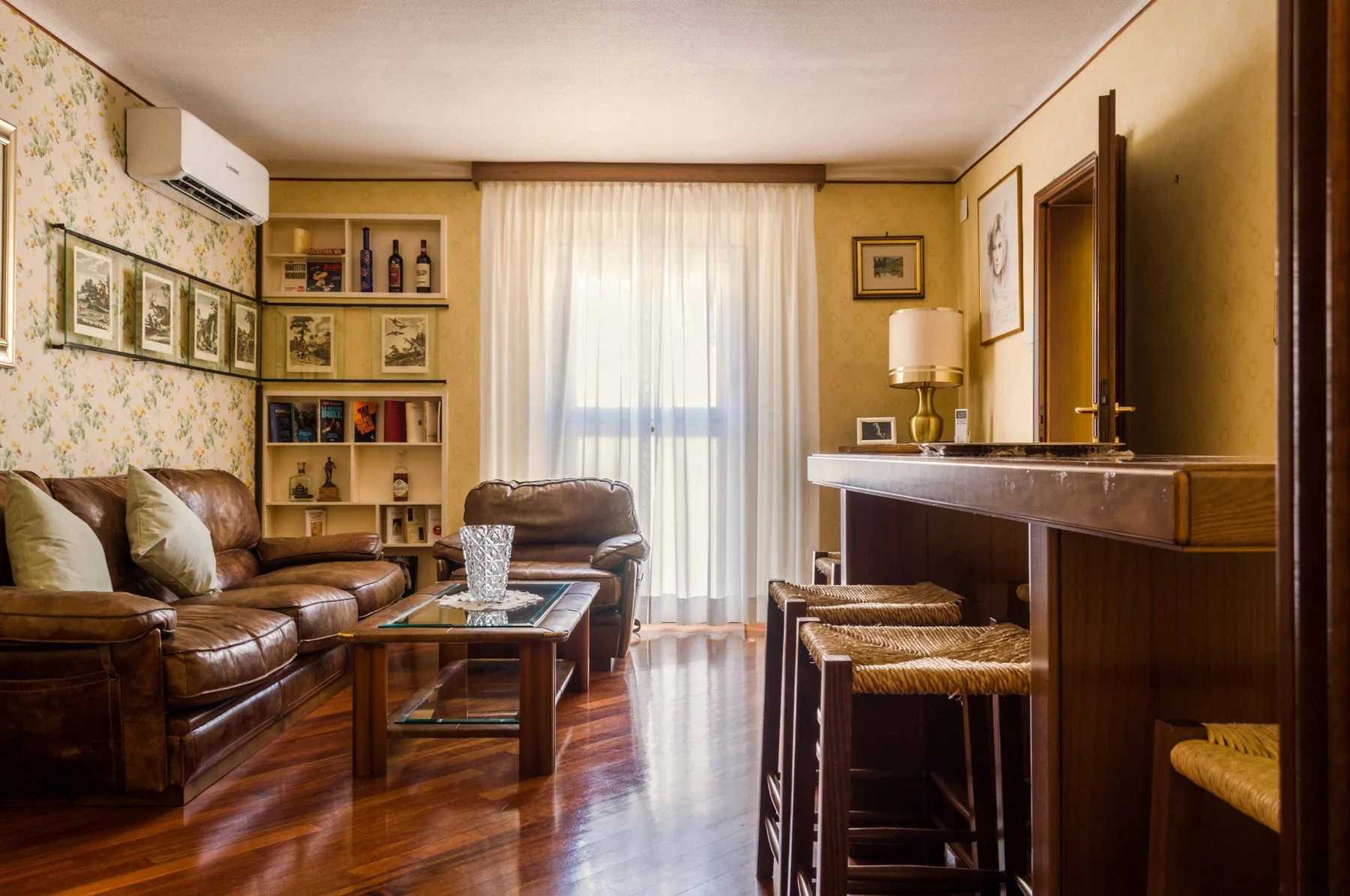 A living room with brown seating, a bookcase, and a bright curtained window.