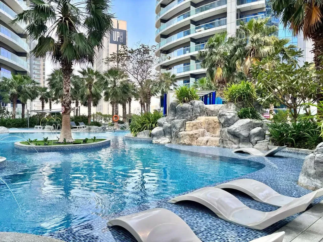 An outdoor pool with curved in-water loungers, palm trees, and modern buildings.