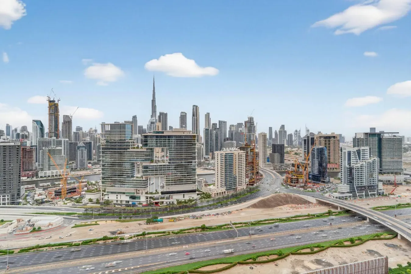 A city skyline rises above multilane roads beneath a blue, cloud-dotted sky.