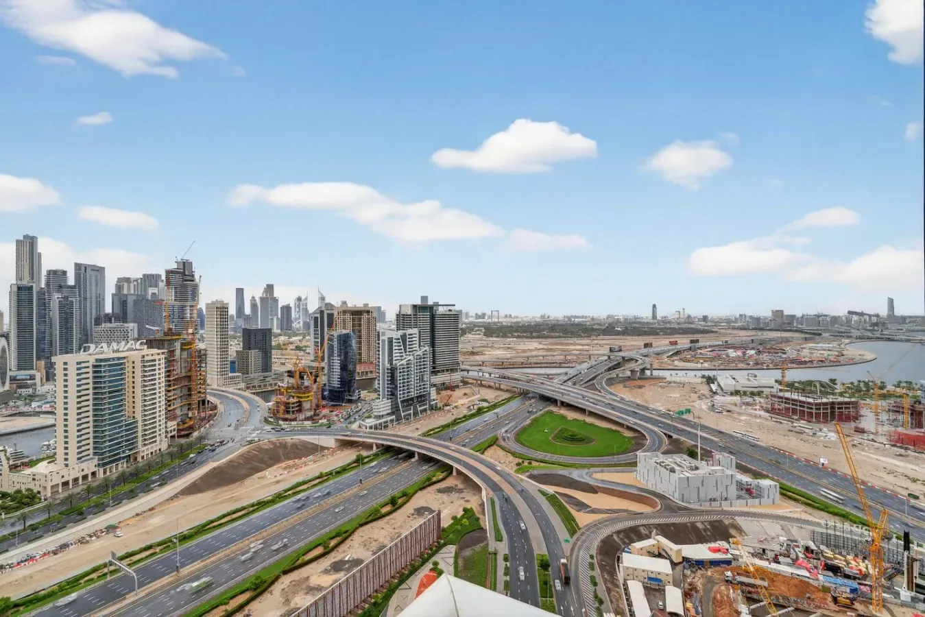 Aerial cityscape with high-rise buildings, curving roads, and construction sites beneath a blue sky.