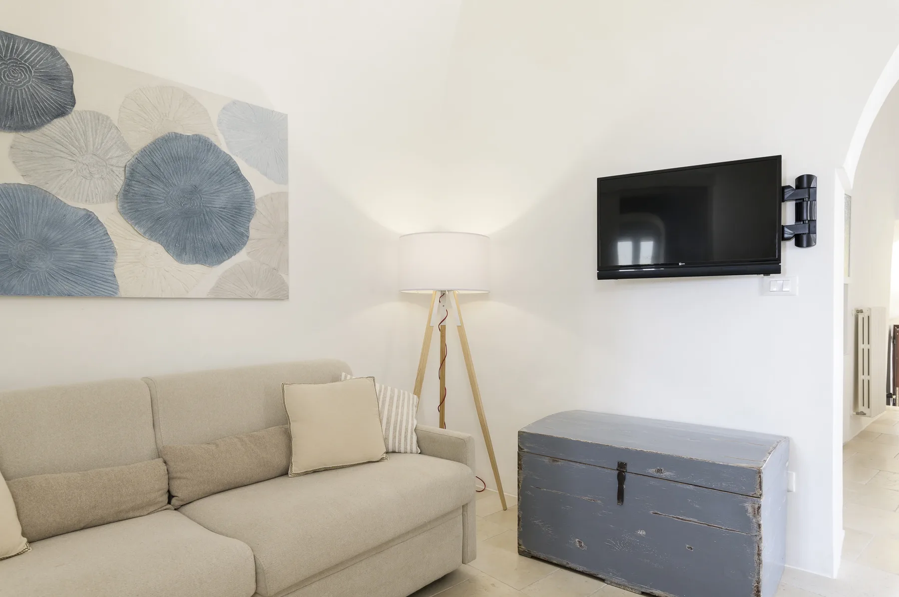 Sala de estar con asientos beige, una lámpara de pie y un televisor en la pared.