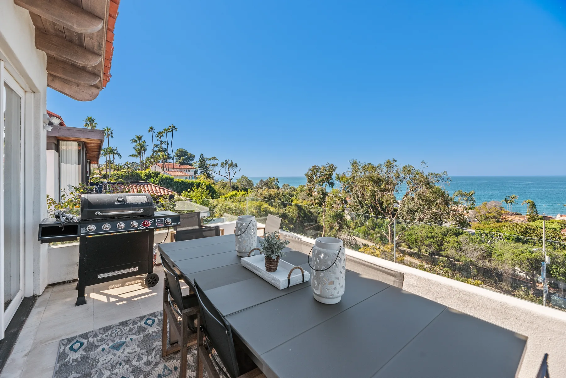 Una mesa de comedor al aire libre y una parrilla están en una terraza con vistas a árboles y al mar.