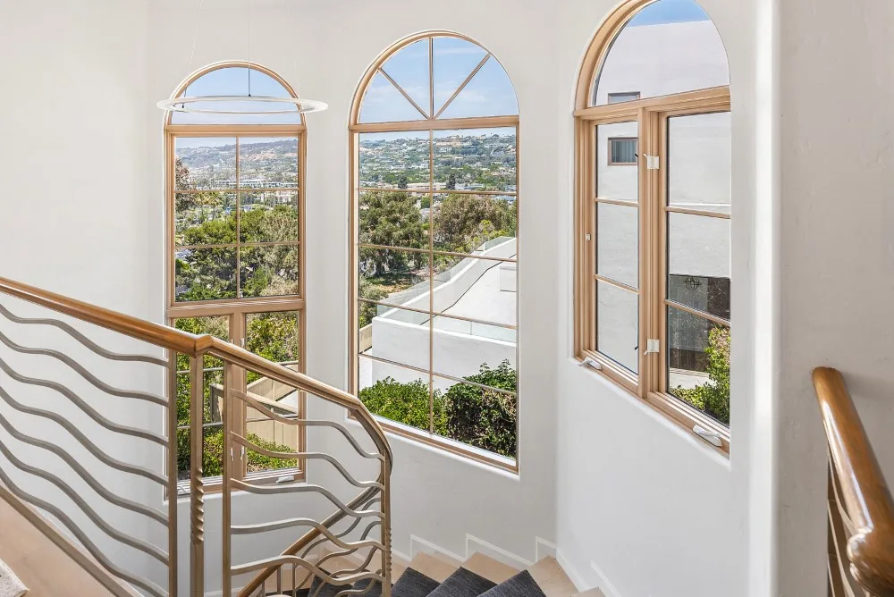 Una escalera luminosa con ventanas altas en arco y vistas a árboles y edificios cercanos.