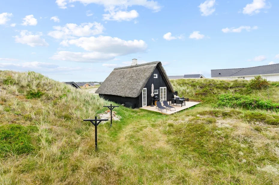 A thatched cottage sits among grassy dunes with outdoor seating by the entrance.
