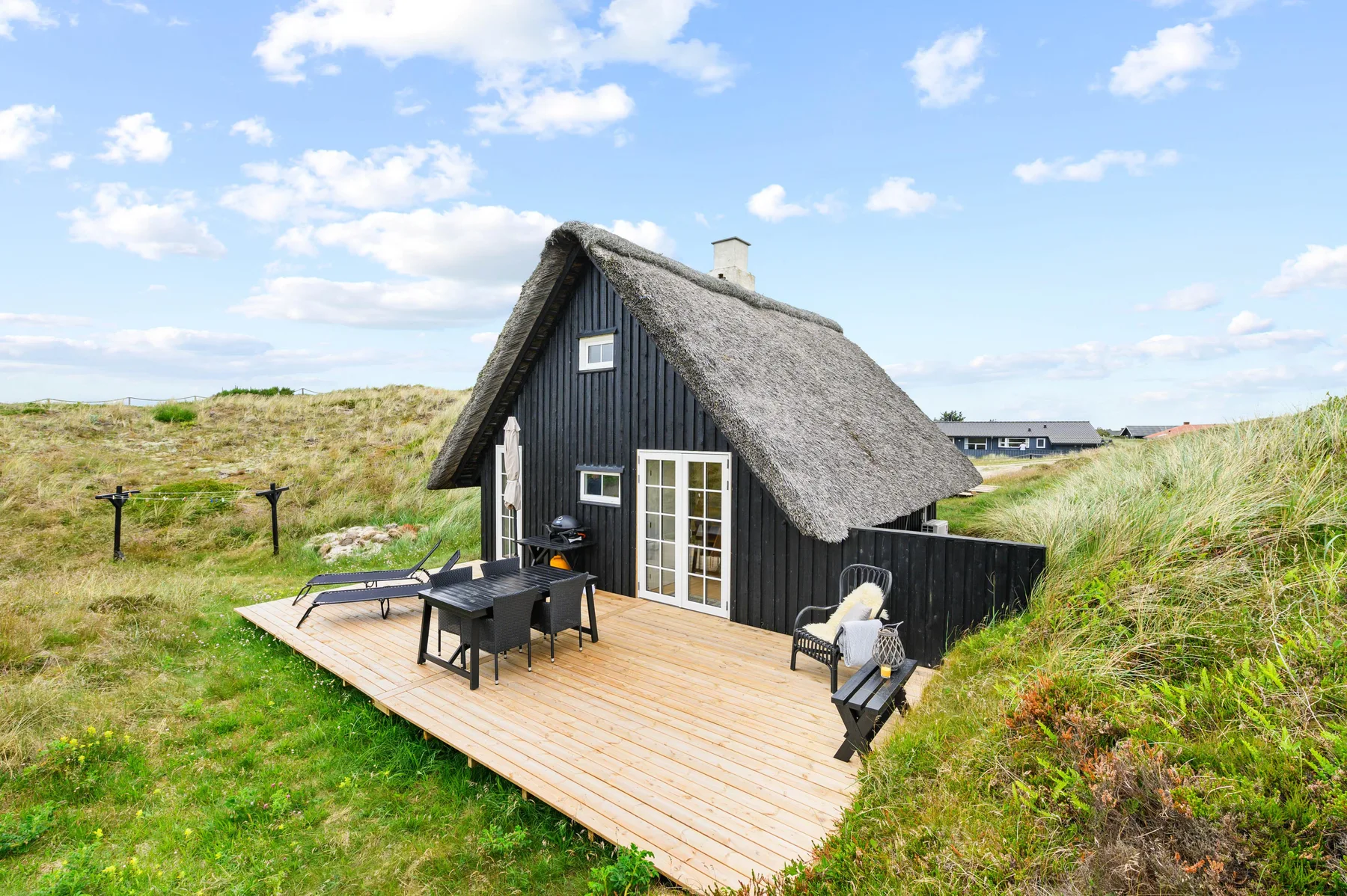 A dark cottage with a thatched roof and wooden deck sits among grassy dunes.