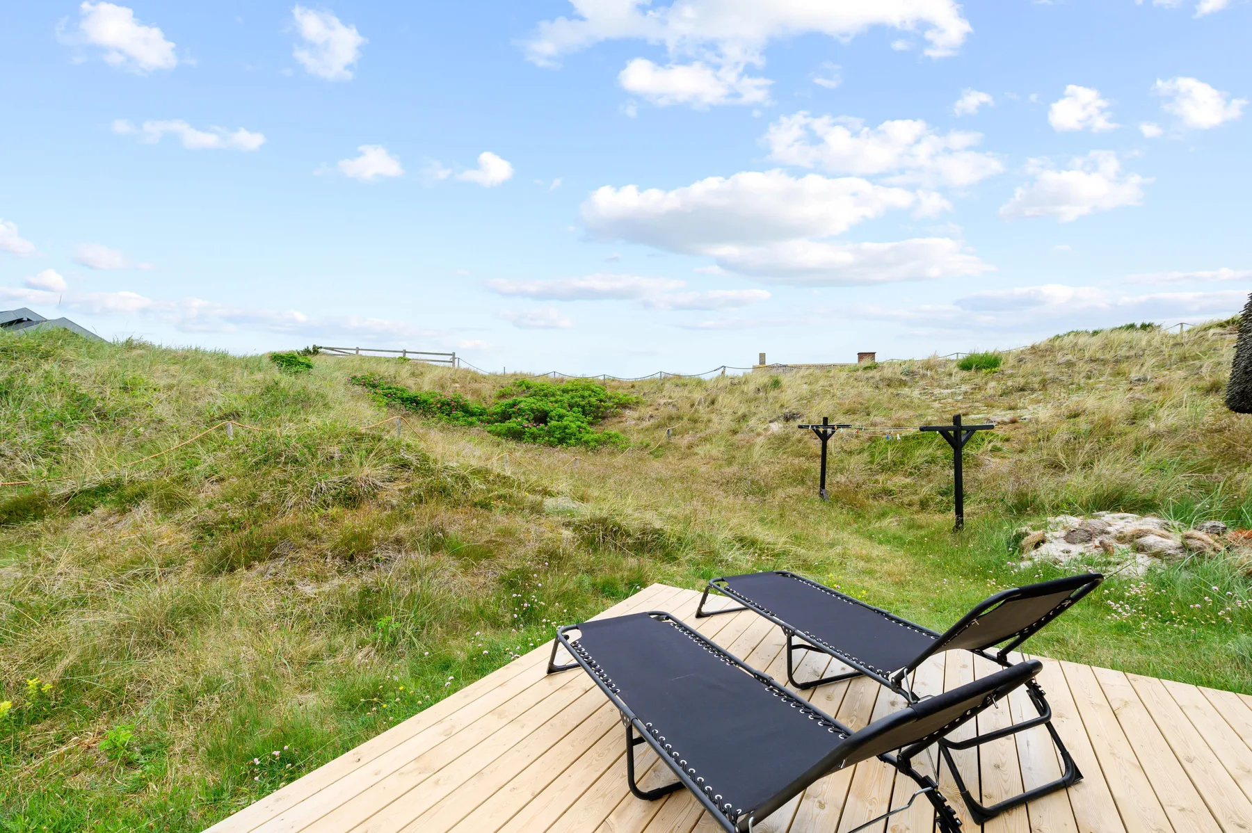 Outdoor loungers sit on a wooden deck beside grassy dunes under a blue sky.