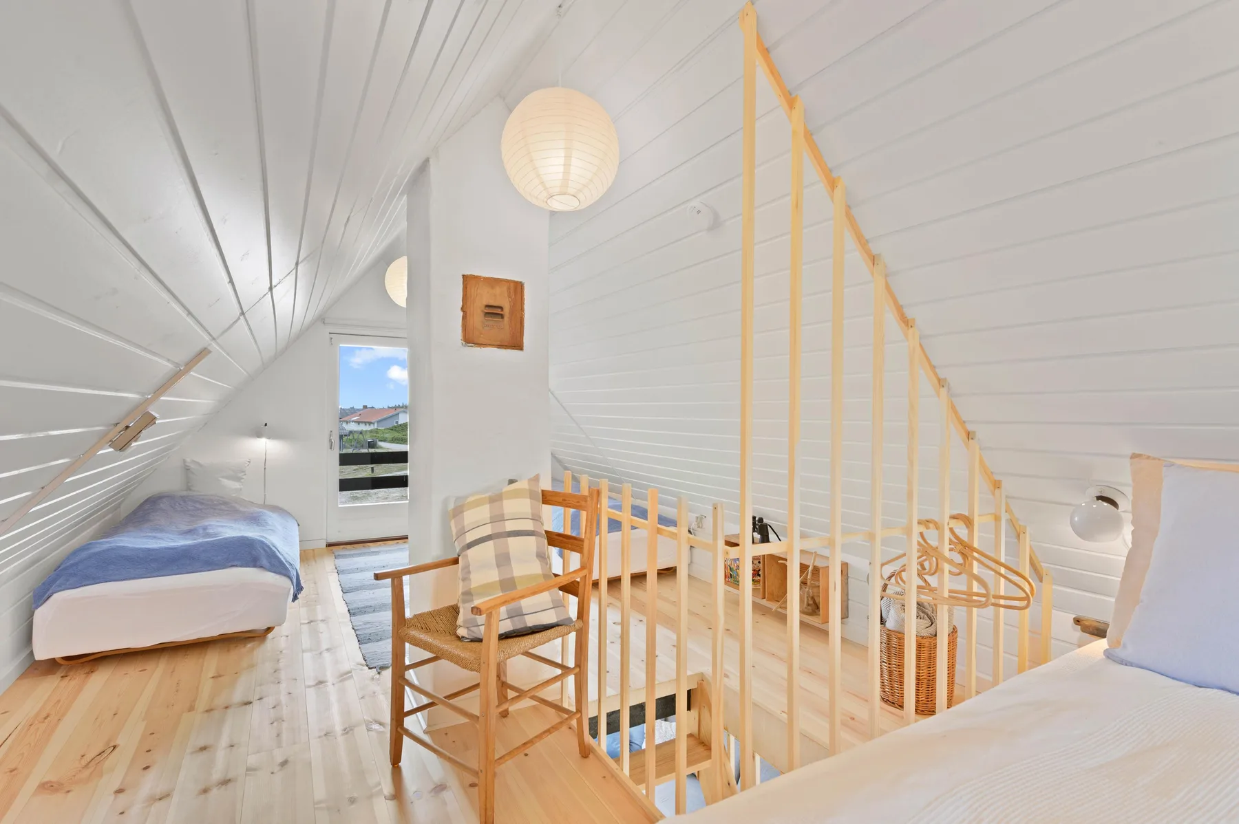 A bright attic bedroom has sloped white ceilings, beds, and a wooden stair railing.