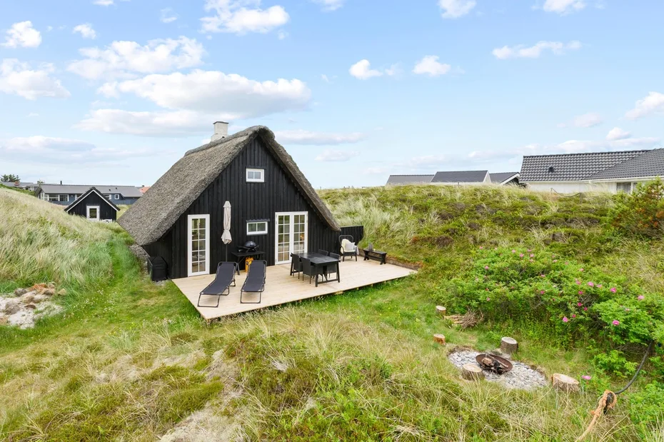 Una casa con techo de paja se encuentra entre dunas cubiertas de hierba, junto a una zona exterior con asientos de madera.