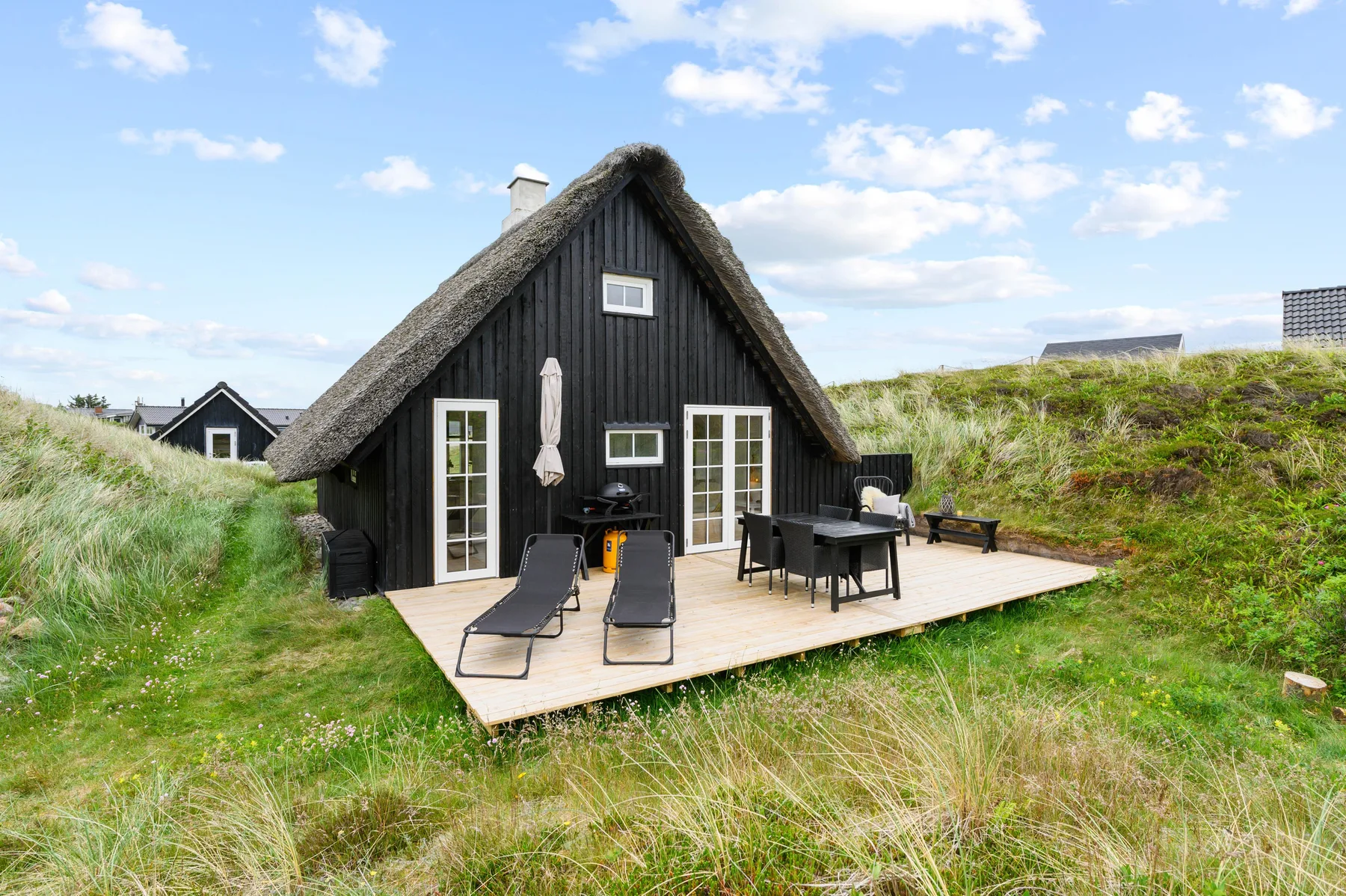 A dark cottage with a thatched roof opens onto a wooden deck with outdoor seating.