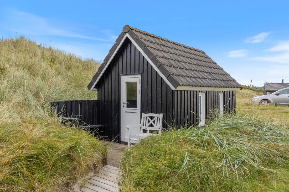 A small dark cottage stands among tall coastal grasses beneath a clear sky.